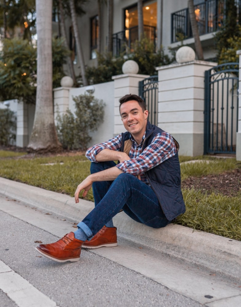 Image of social media influencer Alessandro Pontes sitting outside wearing the Sentinel Plain Toe Boot in Cognac.