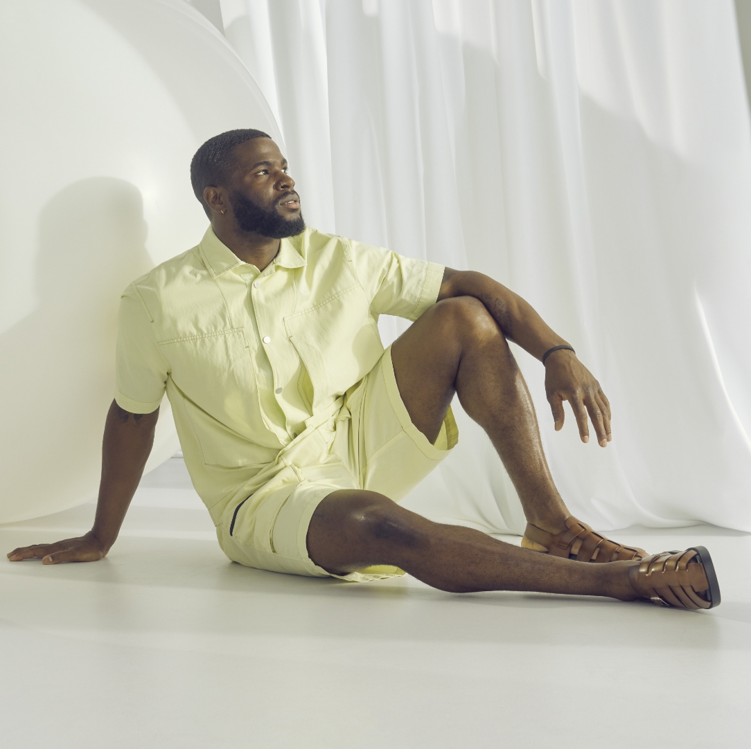 "Finding Men's Fashion Advice Using Social Media." The featured image is a model wearing a matching yellow button down shirt and shorts and brown Stacy Adams sandals while sitting on a white floor with flowing white fabric in the background. 