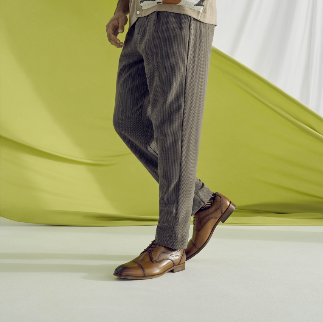 "What Is Business Casual? We're Here To Explain The New Rules." The featured image is a model standing on a white floor with green flowing fabric in the background while wearing dark gray pants and brown Stacy Adams shoes.